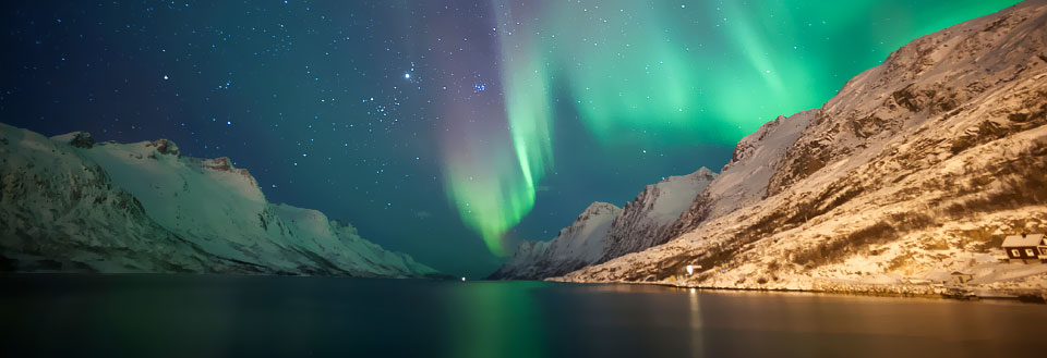 Nordlys stråler over snødekte fjell og et stille vann med et lite hus ved kanten.