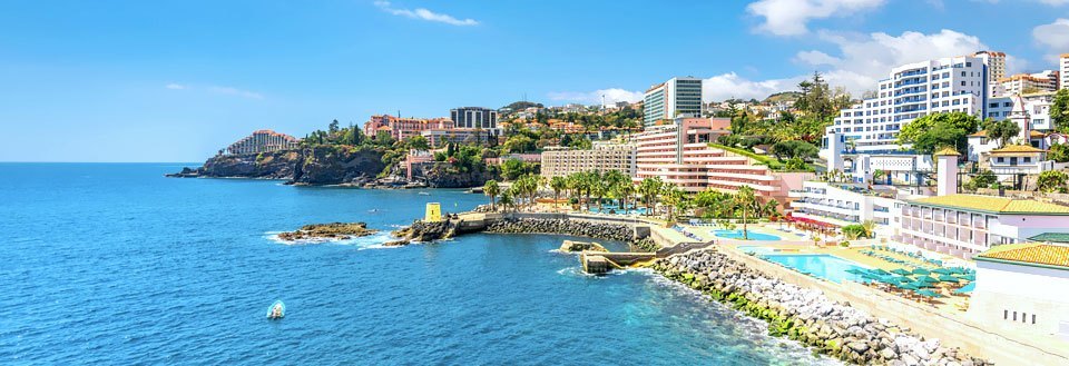 Panoramabilde av Funchal med moderne bygninger, hoteller og et sjøbasseng langs strandlinjen under en klar blå himmel.