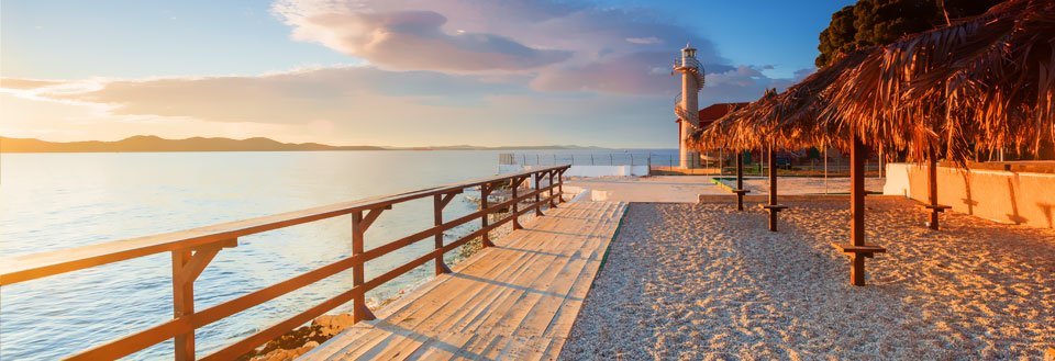 Strandpromenade under solnedgang med et fyrtårn, en trebro og palmebladhytter.