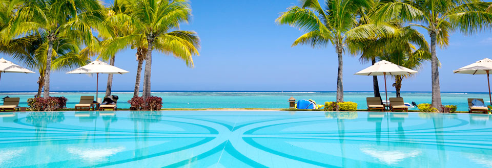 Luksuriøst feriested med svømmebasseng foran en tropisk strand med palmetrær og parasoller.