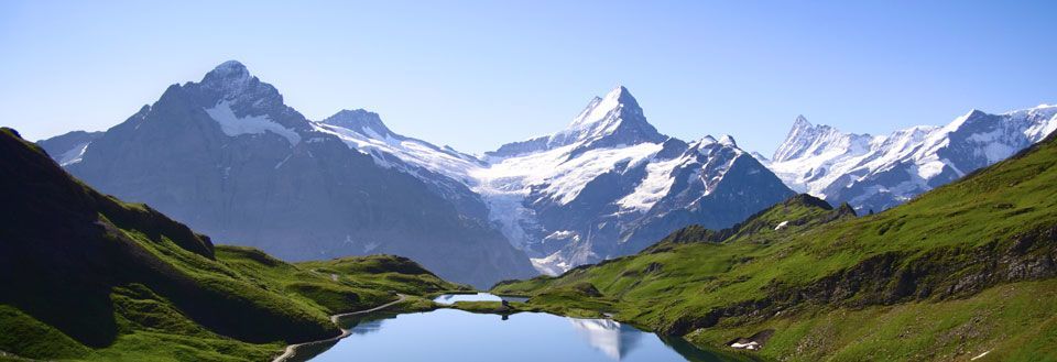 Idyllisk fjellandskap med en krystallklar blå innsjø i forgrunnen og snødekte topper i bakgrunnen.