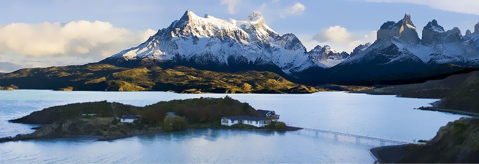 Fjellandskap med snødekkede topper, en innsjø i forgrunnen, og spredte bygninger under en skyet himmel.