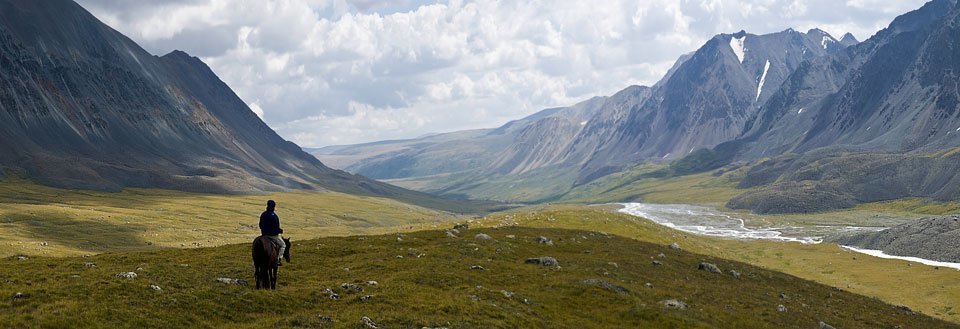 En rytter på en hest betrakter et majestetisk fjellandskap med vide sletter og en elv.