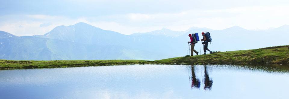 To fjellvandrere med sekker går ved en fjellsjø, omgitt av majestetiske fjell.